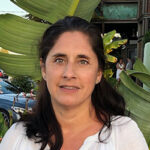 A woman with long dark hair and a white top stands outdoors in front of large green leaves. There are people and a building in the background.