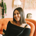 A young woman with straight brown hair, wearing a black off-the-shoulder top, sits and smiles on an orange couch. There is a blurred figure and a cactus in the softly lit background.