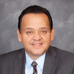 A man with dark hair wearing a gray suit, light blue shirt, and patterned tie, smiling in front of a gray studio background.