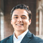 Paul Flores: A young man with dark hair, wearing a dark suit jacket and white dress shirt, smiles confidently while standing in a corridor with blurred brick and stone details in the background.